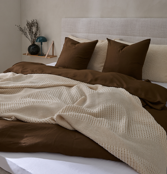 Redt seng med brunt dynebetræk, beige strikket plaid og matchende puder i et neutralt soveværelse med polstret sengegavl og lille hylde med vase og lampe.