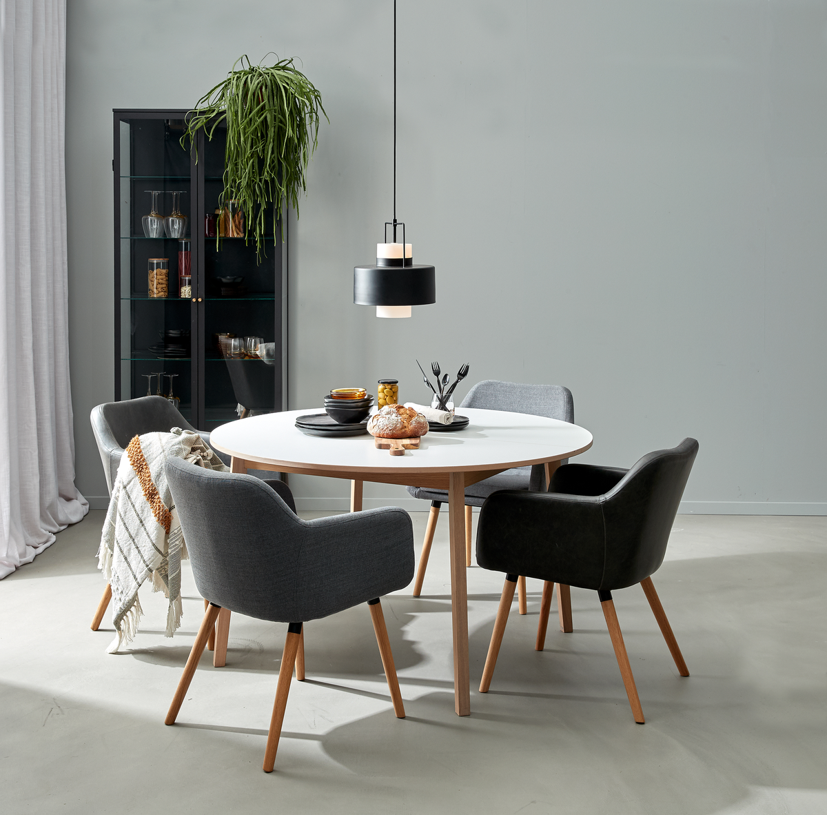Rundt hvidt spisebord med træben og fire polstrede stole under en sort pendellampe; vitrineskab, hængende plante og plaid i en minimalistisk spisestue.