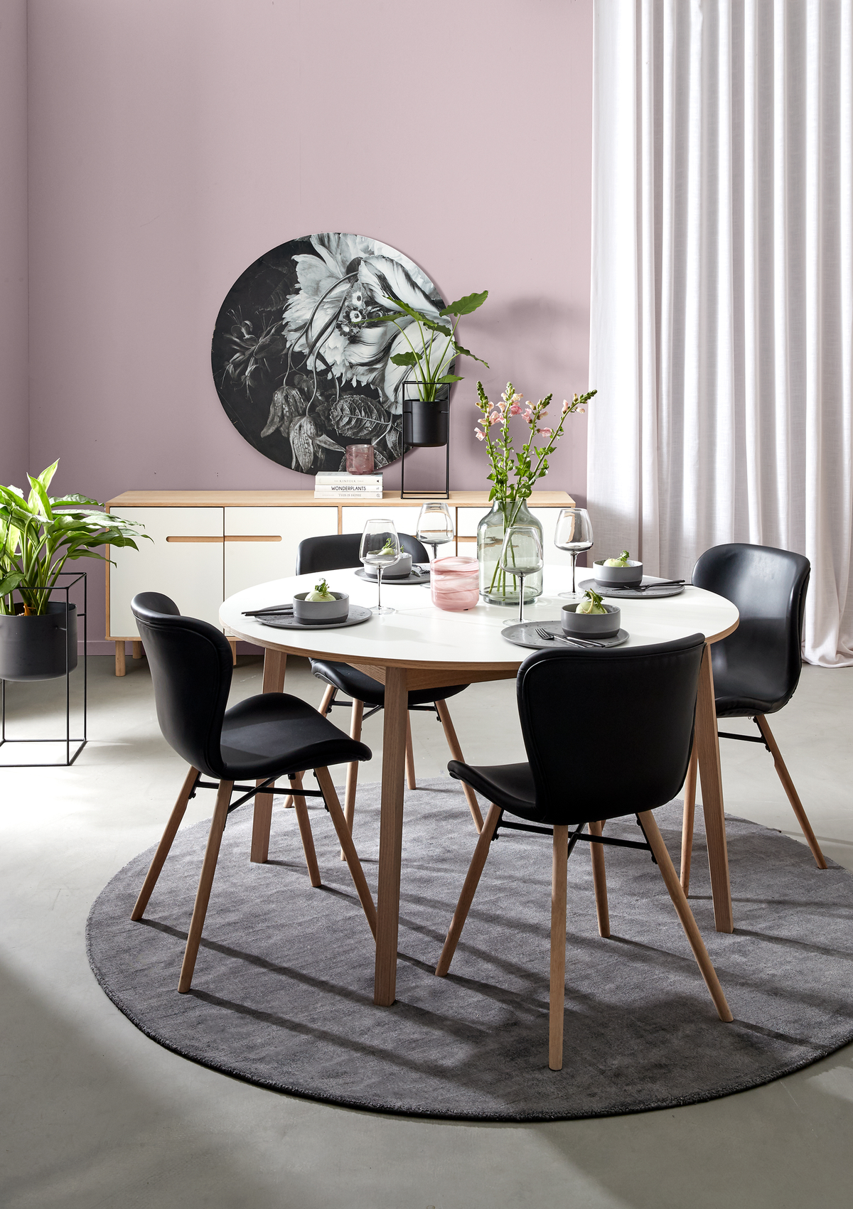 Moderne spiseplads med rundt hvidt bord, sorte stole med træben og rund grå måtte; grønne planter, glasvase med blomster og opdækning med vinglas foran skænk med rund vægdekoration og lyse gardiner i dagslys.