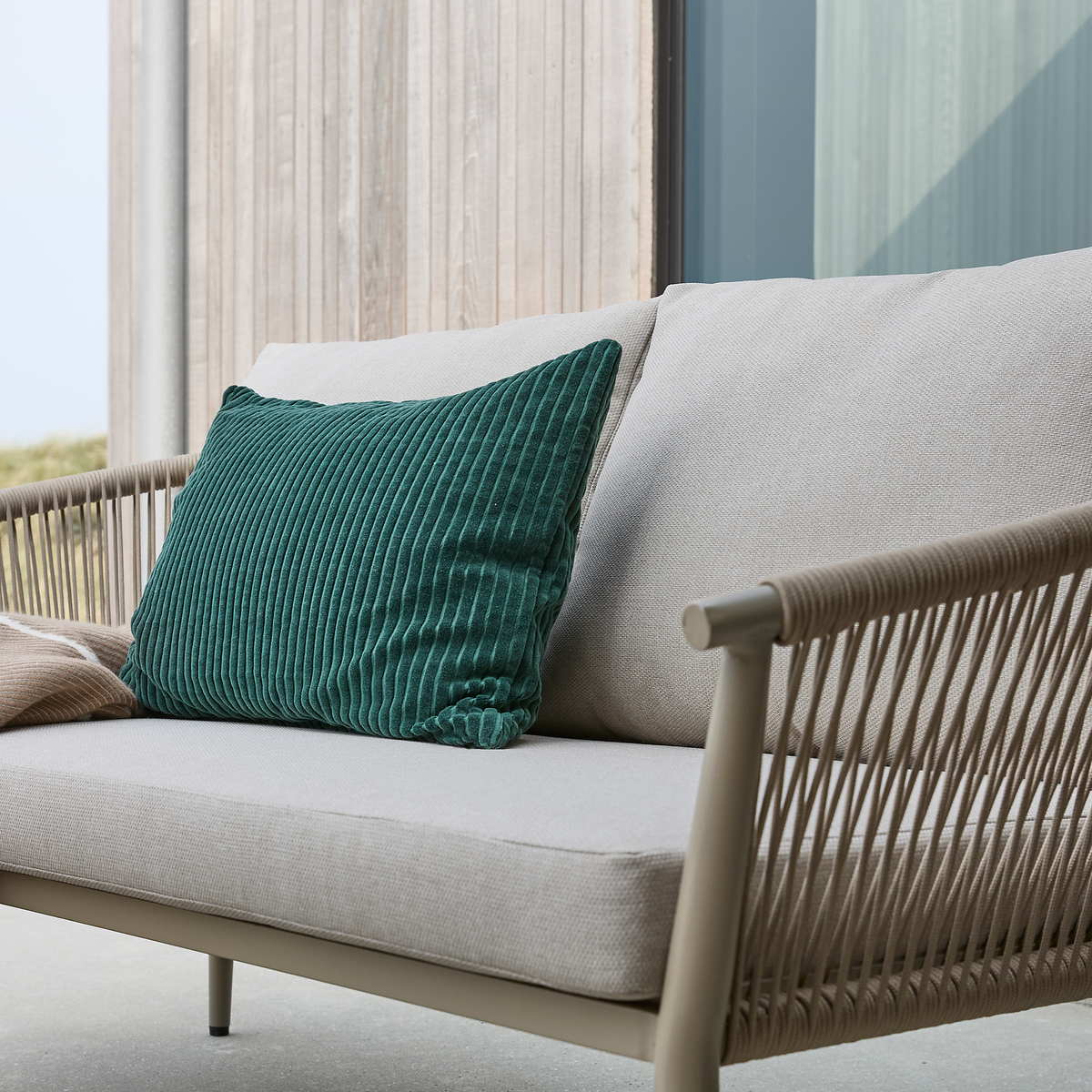 Beige udendørs sofa med riflet grøn pude og beige plaid ved en træfacade.