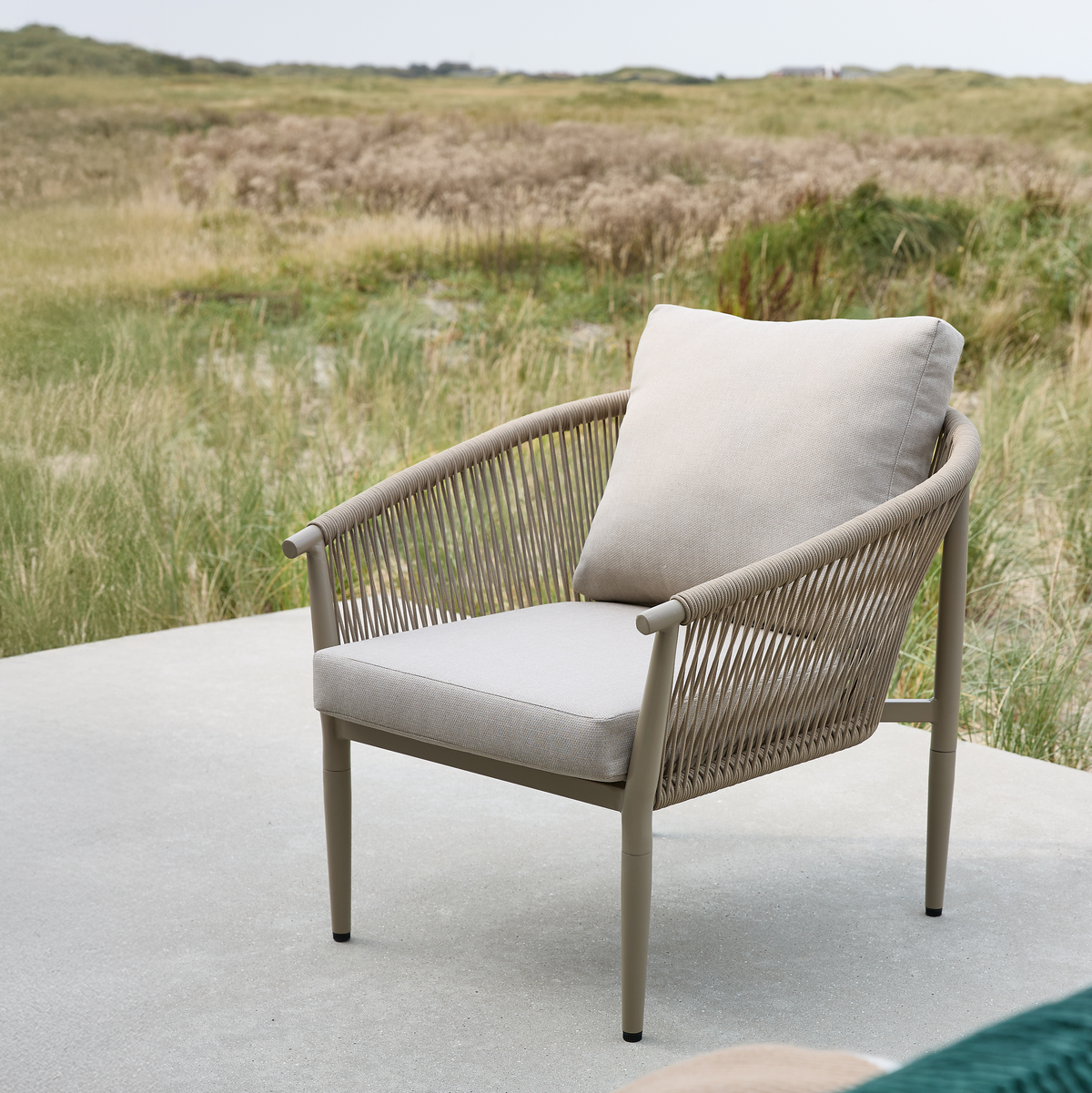 Lys beige udendørs lænestol med rebdetaljer og bløde hynder på en betonterrasse med græsklædte klitter i baggrunden.