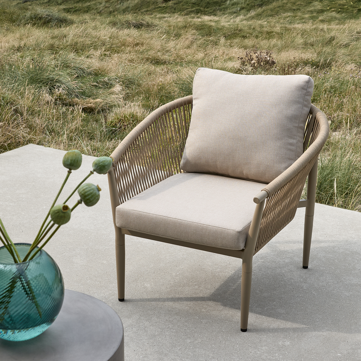 Beige udendørs lænestol med hynder på betonterrasse, ved siden af turkis glasvase med grønne frøkapsler; græsklædt natur i baggrunden.
