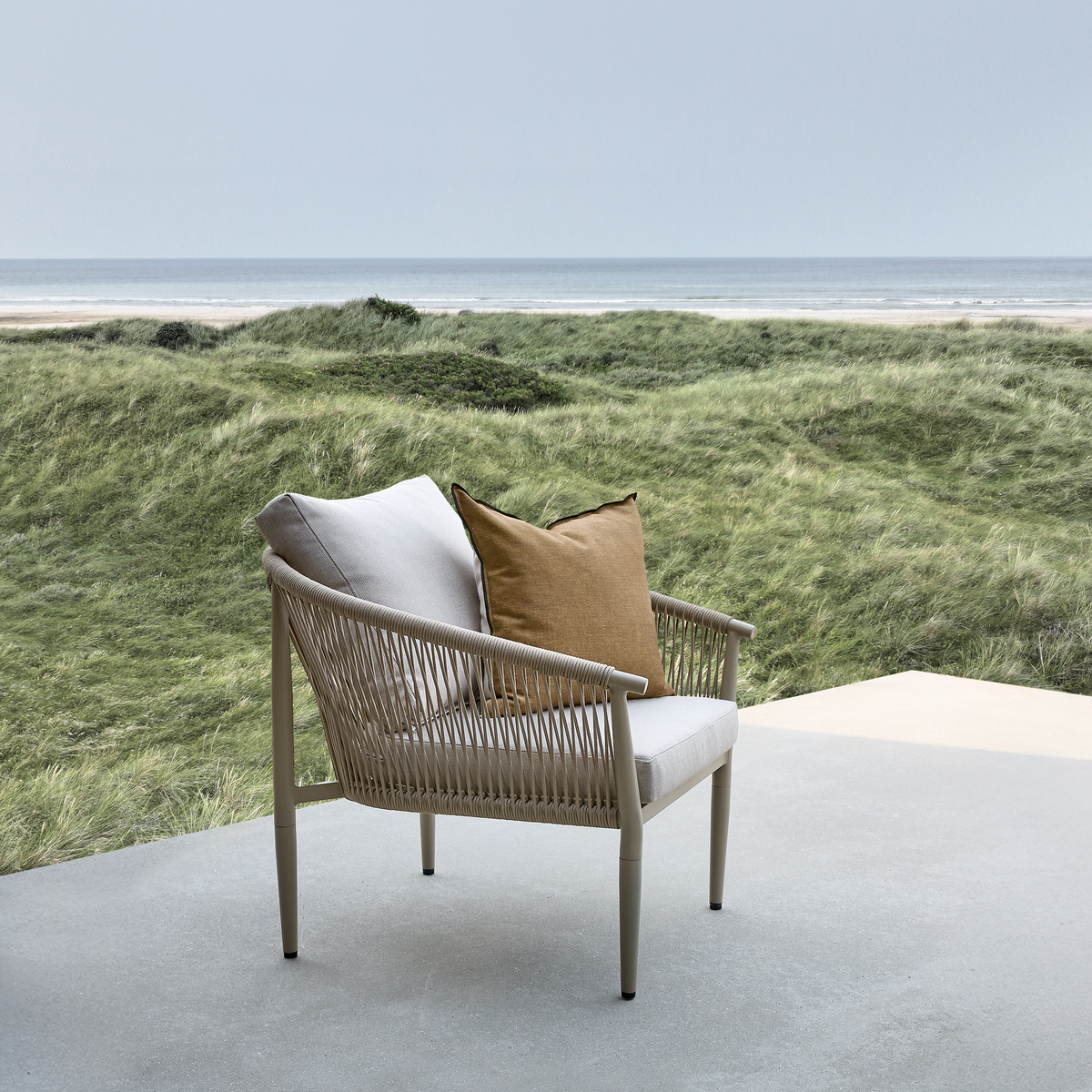 Udendørs lænestol med beige stel og flettede sider, lysgrå hynde og okkerfarvet pude på en terrasse med udsigt over græsklædte klitter og havet i dagslys.