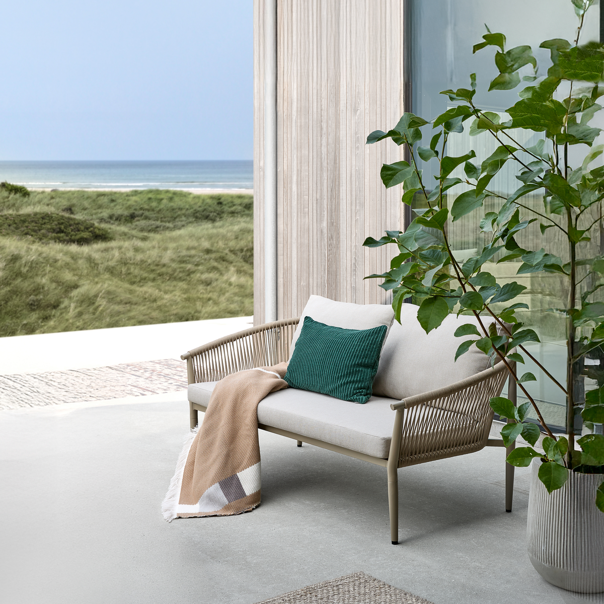 Udendørs bænk med beige hynder, grøn pude og plaid på en terrasse med udsigt til hav og klitter; stor potteplante ved siden af.