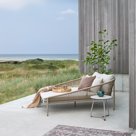 Terrasse med loungemøbel med puder og plaid, lille sidebord med glasvase, grønt klitlandskab og hav i baggrunden ved et hus med træbeklædning.