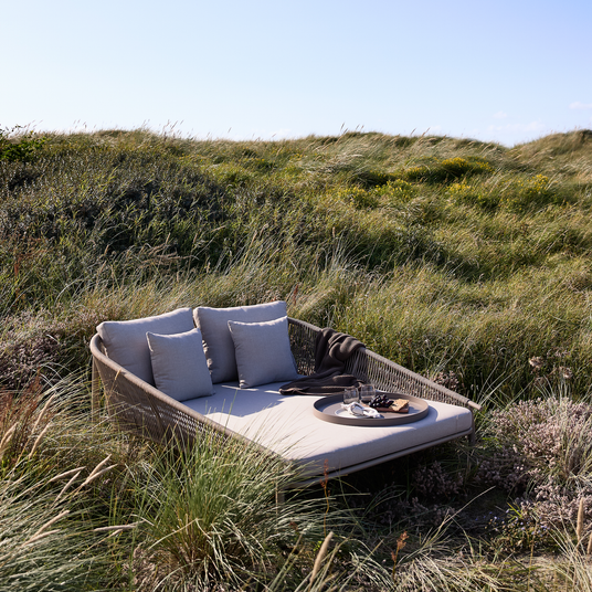 Lys udendørs daybed med beige puder og en bakke med glas og druer, placeret i klitlandskab under blå himmel.