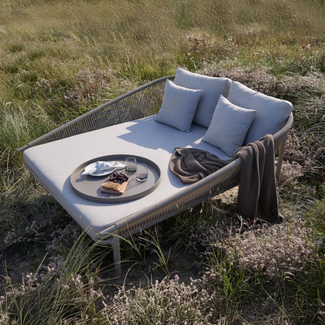 Udendørs daybed med lysegrå hynder, puder og plaid; bakke med glas, serviet, kiks og druer i en græsmark i dagslys.