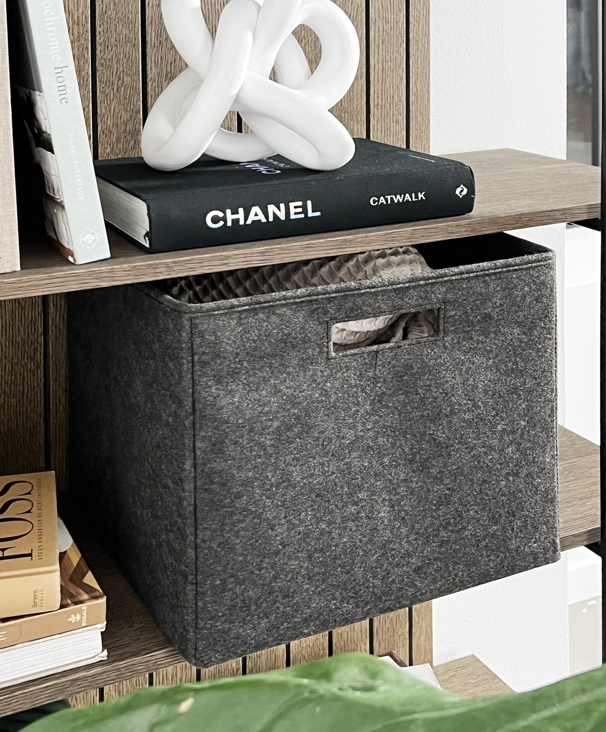 Grå filtkasse på en træhylde ved siden af bøger, bl.a. CHANEL Catwalk, med en hvid keramisk knudeskulptur ovenpå.