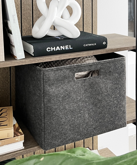Grå filtkasse på en træhylde ved siden af bøger, bl.a. CHANEL Catwalk, med en hvid keramisk knudeskulptur ovenpå.