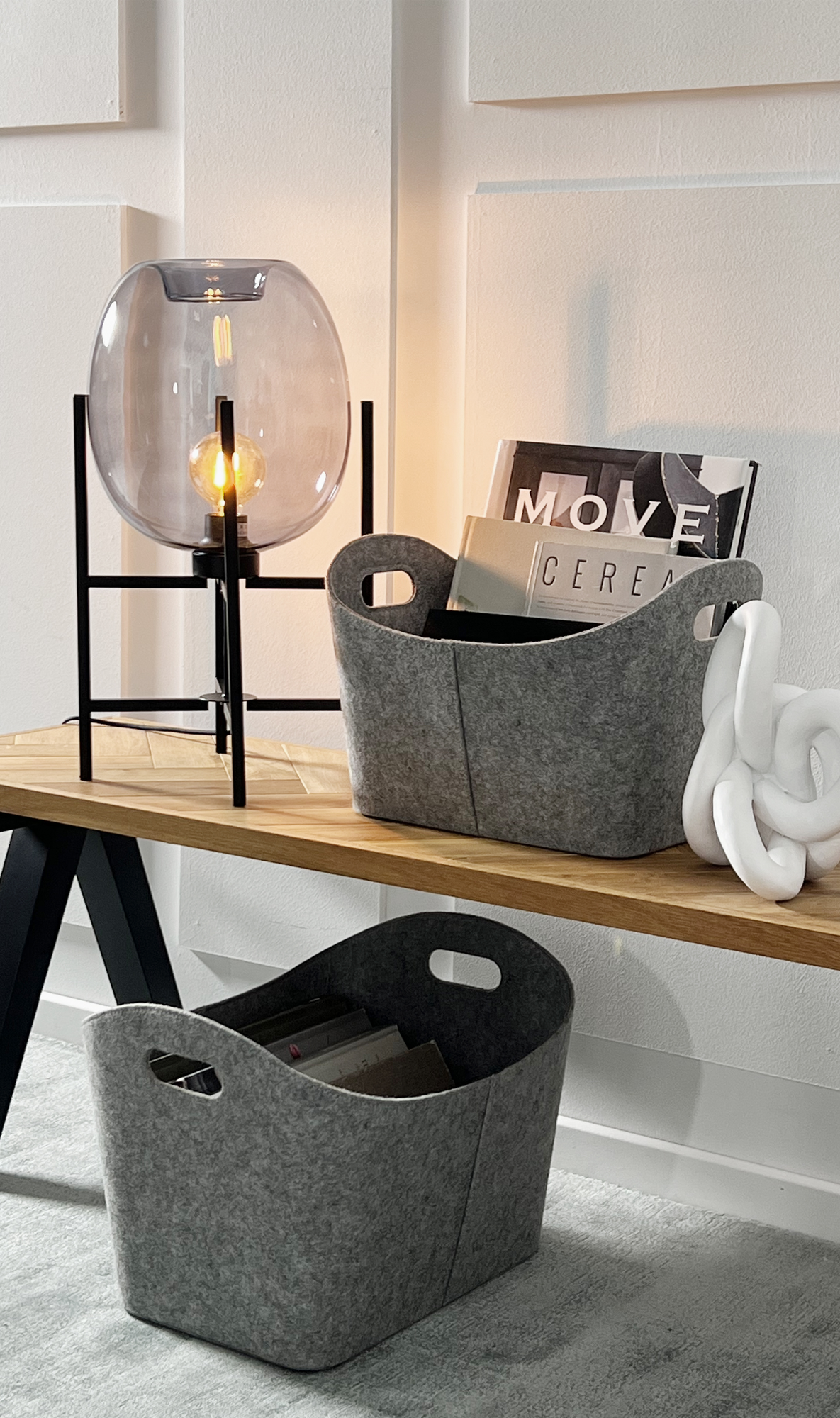 Moderne interiørscene med en bordlampe med glasglob i sort stativ ved siden af en grå filt-kurv med magasiner med titlerne “MOVE” og “CEREA”, en træbænk og en hvid knude-skulptur; endnu en grå kurv står på gulvet.