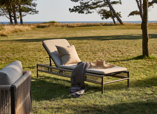 Solseng med beige hynder på græs ved havet, med pude, plaid og stråhat i varm aftensol.