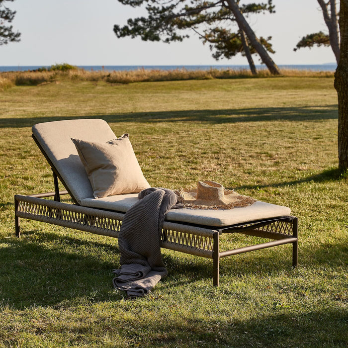 Solseng med beige hynder på græs ved havet, med pude, plaid og stråhat i varm aftensol.