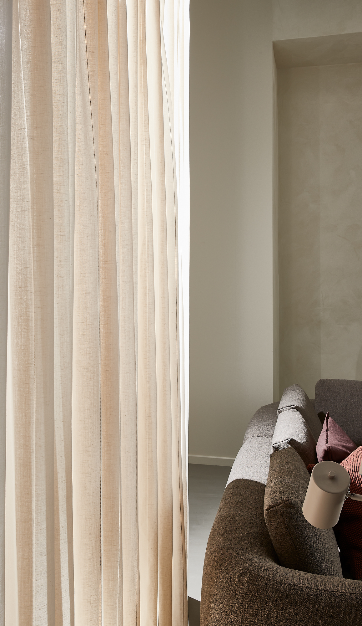 Lyse beige, lette gardiner ved siden af en gråbrun sofa med rosa puder og en lille beige læselampe i blødt dagslys.