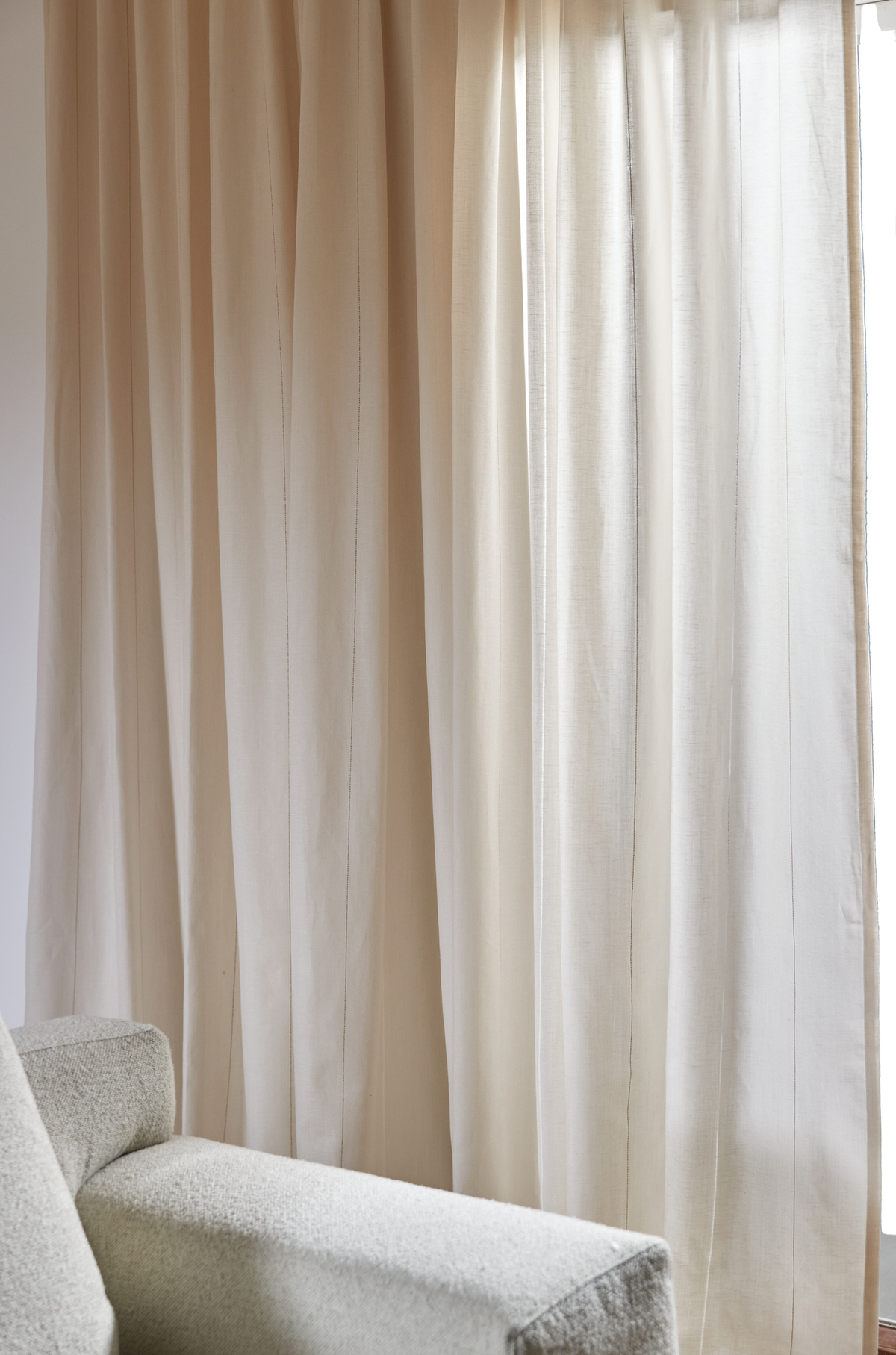 Beige, let transparente gardiner foran et vindue i dagslys, med et gråt sofa-armlæn i forgrunden.