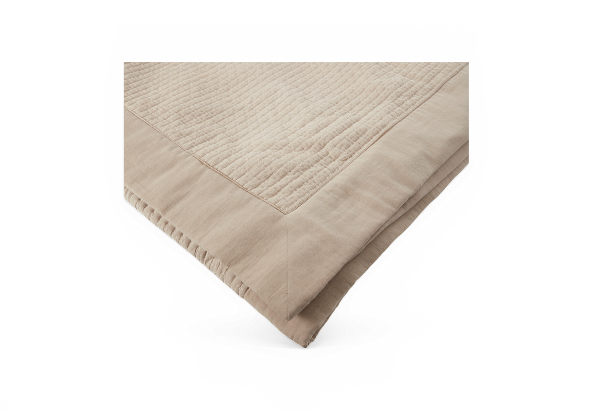 Beige quiltet sengetæppe, foldet med hjørnedetalje.