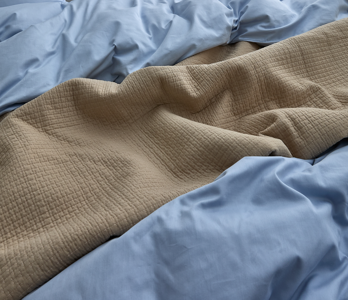 Beige quilt med kvadratisk quiltning mellem krøllede lyseblå lagner i nærbillede.