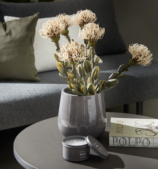 Grå vase med lyse blomster på rundt sofabord; duftlys MOOD i grå dåse og bogen POLPO ved siden af, sofa med puder i baggrunden.