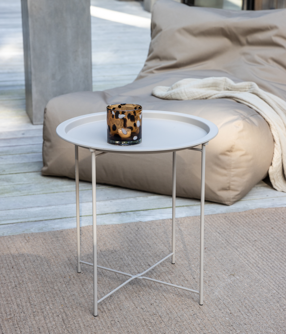 Lille rundt sidebord i lysegrå metal på en træterrasse med en mønstret glaslyslygte; beige sækkestol og lys plaid i baggrunden.