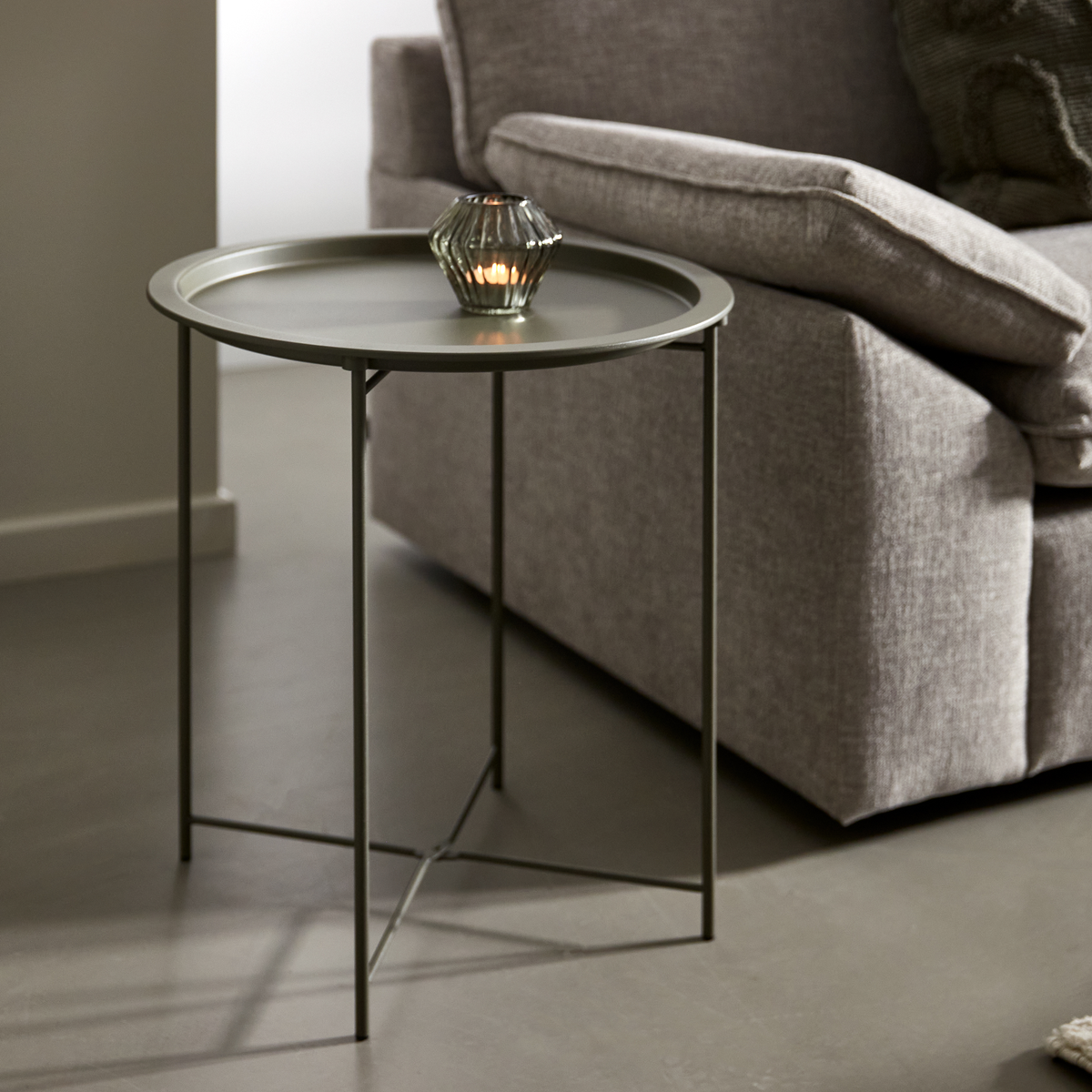 Lille rundt sidebord i olivengrøn metal med glaslysestage med tændt fyrfadslys ved siden af en grå sofa i stuen.