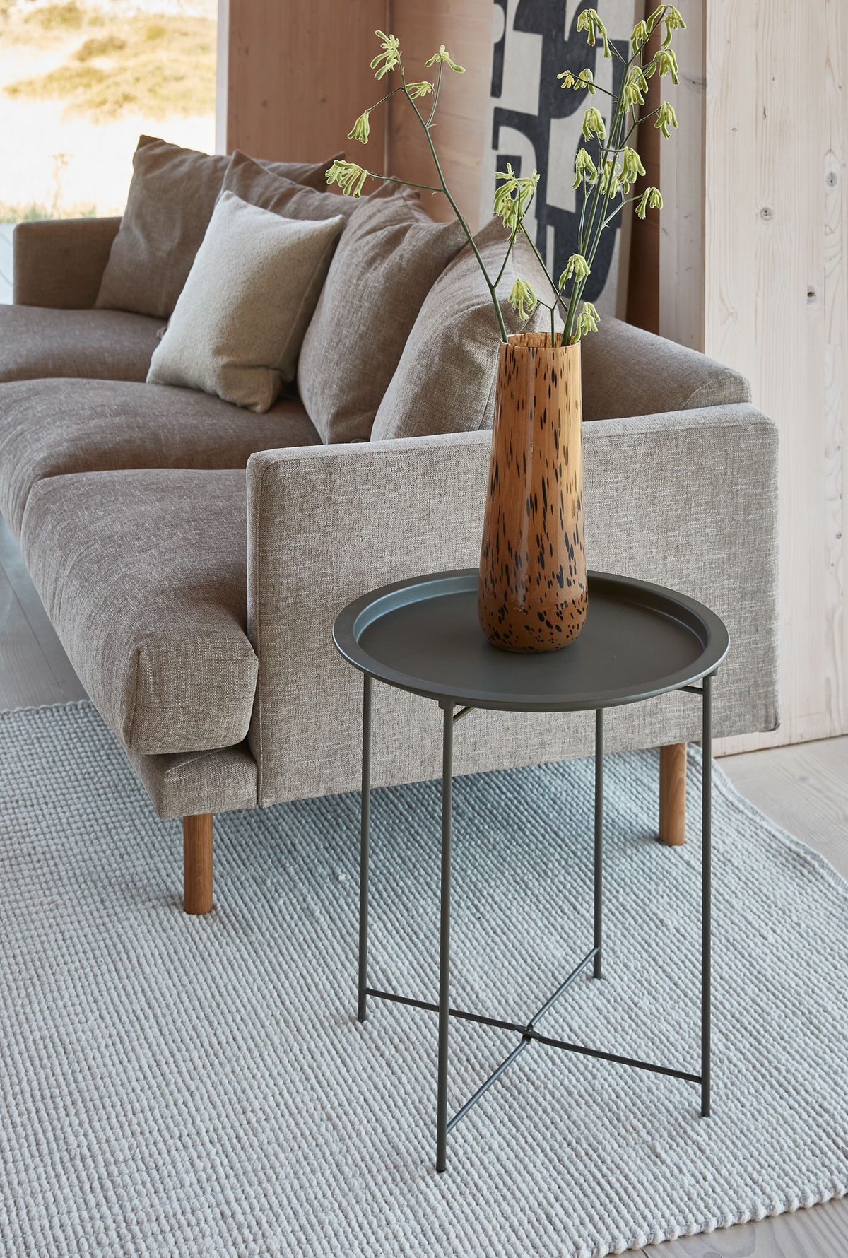 Minimalistisk stue med beige sofa, lys tæppe og rundt metal sidebord med høj trævase med grønne grene.
