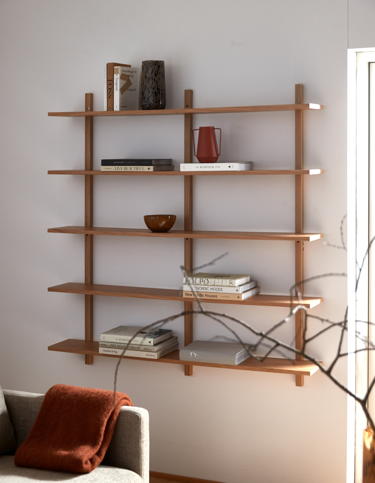 Vægmonteret reol i lyst træ med stabler af bøger – Live Beautiful, Kinfolk, Nordic Moods, New Nordic Houses, Dior – samt keramikvase, rød kande og skål; beige stue med plaid over sofa i blødt dagslys.
