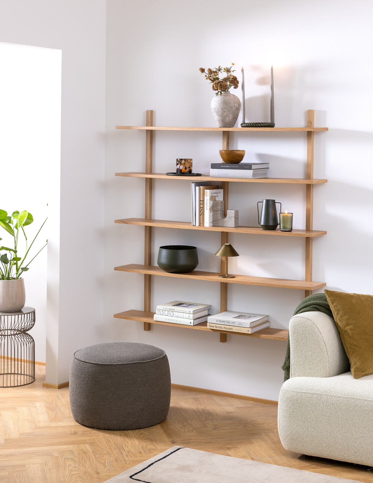 Lyst skandinavisk hjørne i stuen med vægmonteret eg-reol pyntet med vaser, bøger som New Nordic Houses, lille bordlampe og stearinlys; beige sofa, grøn plante og grå puf.