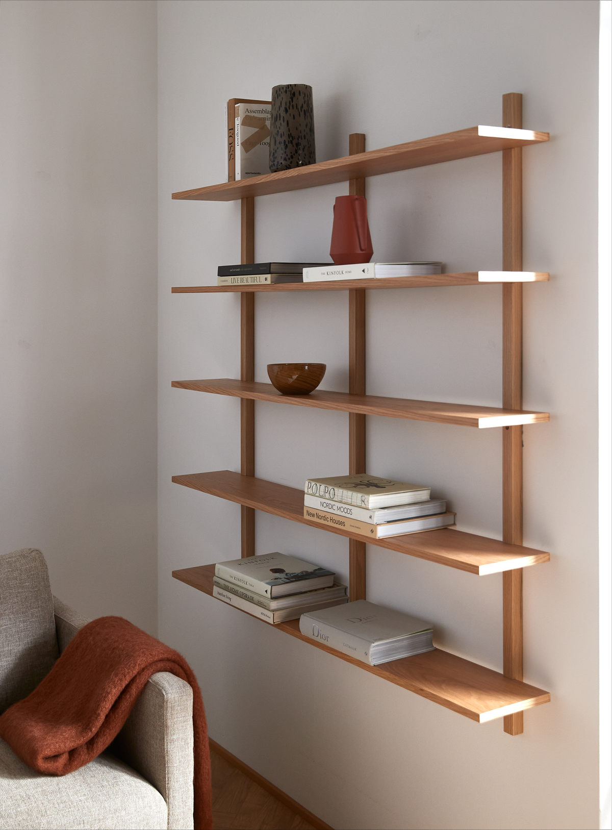 Vægmonteret træreol med bøger som Kinfolk, New Nordic Houses og Dior, keramisk vase, rød kande og lille træskål; beige sofa med rustfarvet plaid ved siden af.