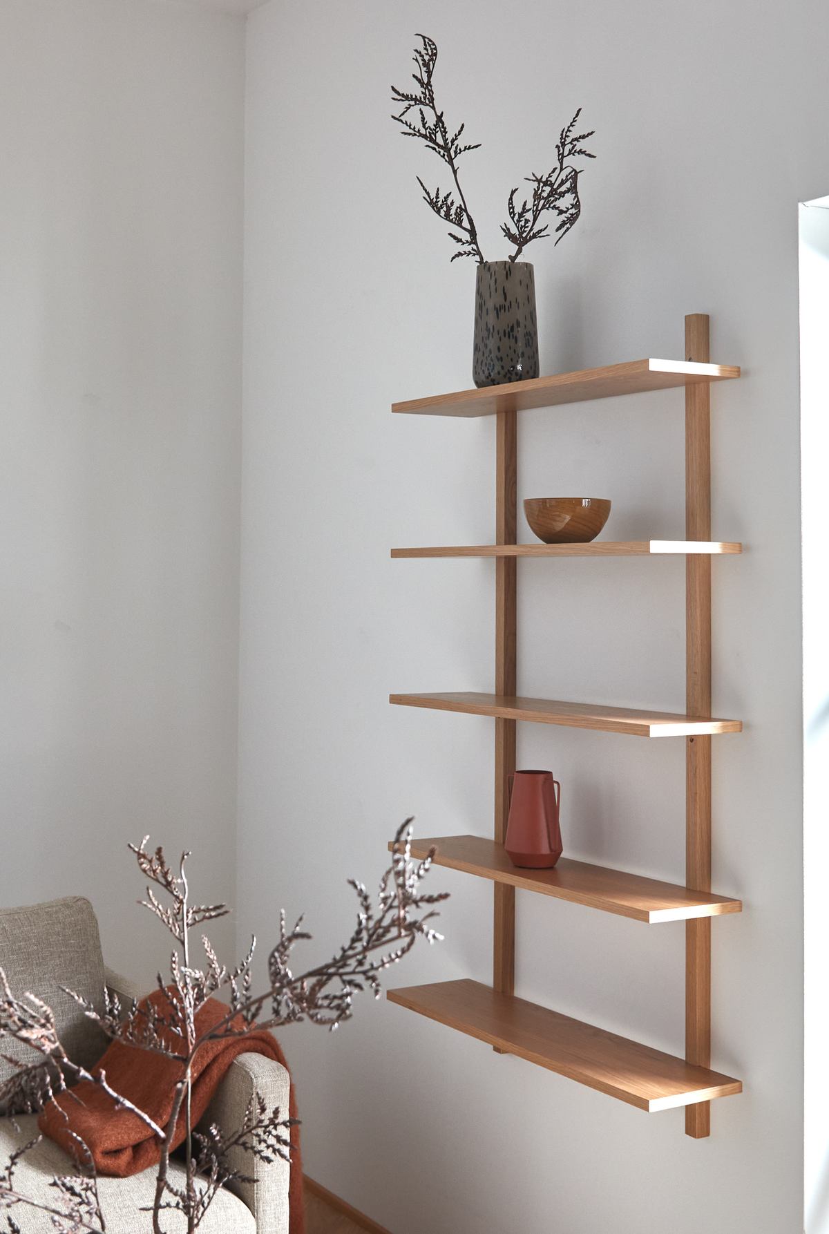 Vægmonteret reol i lyst træ med vase med grene, træskål og terracottakande; beige sofa med rustfarvet plaid i en lys stue.