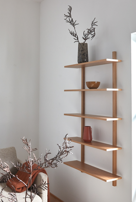 Vægmonteret reol i lyst træ med vase med grene, træskål og terracottakande; beige sofa med rustfarvet plaid i en lys stue.