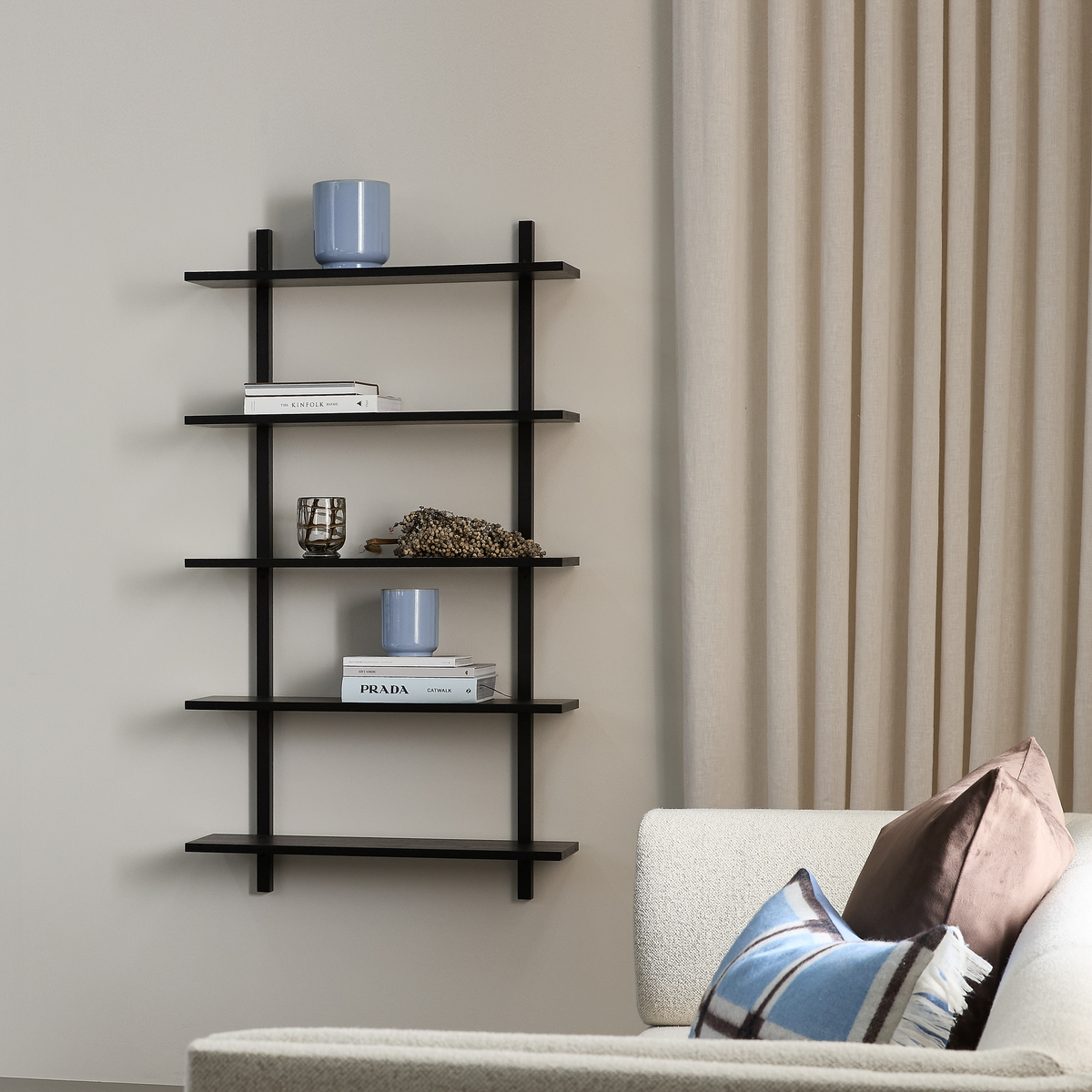 Moderne stue med sort vægmonteret hylde stylet med blå vaser/lys, glas og dekoration; bøgerne Prada og Kinfolk; beige gardiner og lys sofa med blå og brun pude.