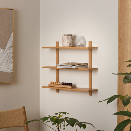 Væghylde i eg med tre hylder, pyntet med vase, krukke, magasiner og træperlekugler; omgivet af grønne planter i en lys stue.