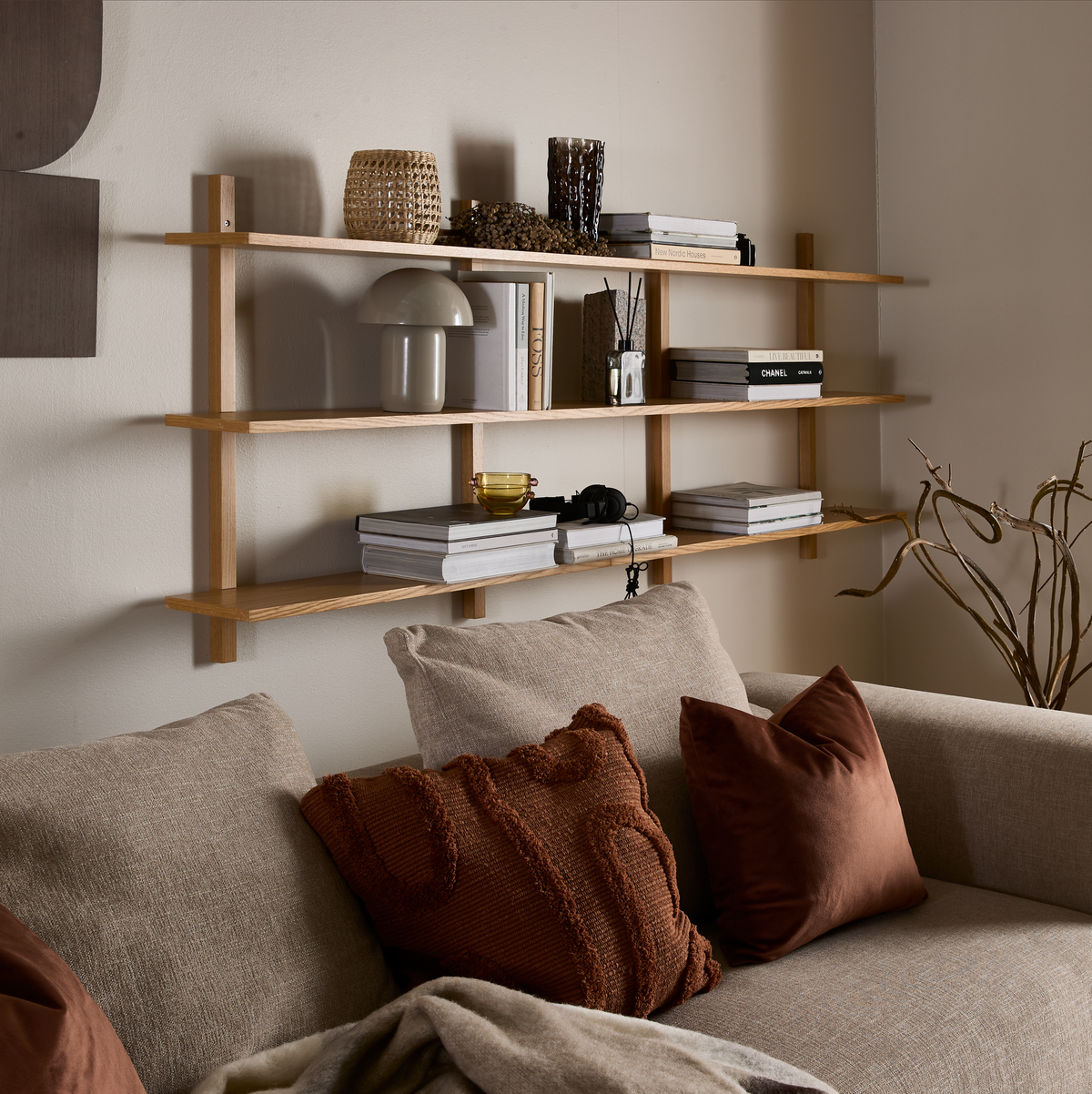Vægmonteret reol i lyst træ over beige sofa med puder; bøger som Chanel og New Nordic Houses, lille bordlampe og flettet kurv på hylderne.