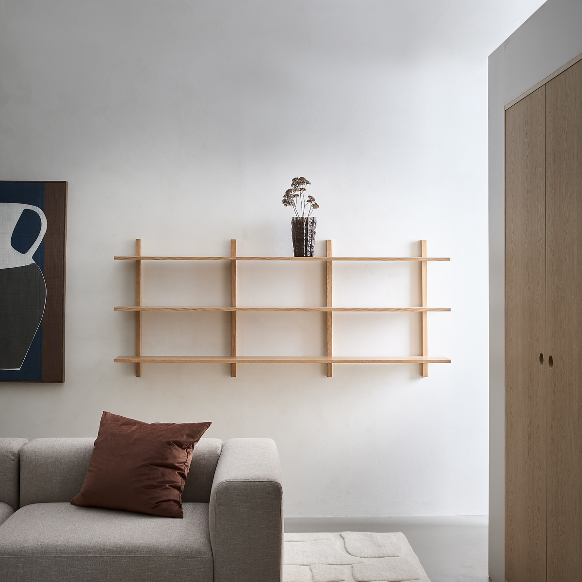 Minimalistisk stue i lyse nuancer med vægmonteret reol i lyst træ, brun glasvase med tørrede blomster, grå sofa med brun pude, abstrakt vægkunst og blød tæppe.