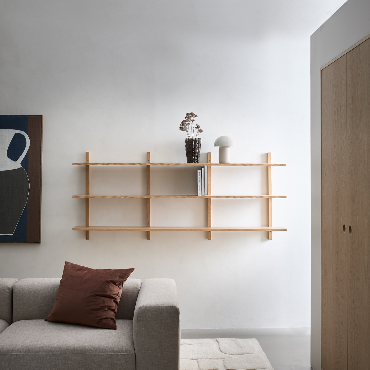 Minimalistisk stue med vægmonteret hylde i lyst træ med vase med tørrede blomster, hvid kuppellampe og et par bøger over en beige sofa og lyst tæppe.