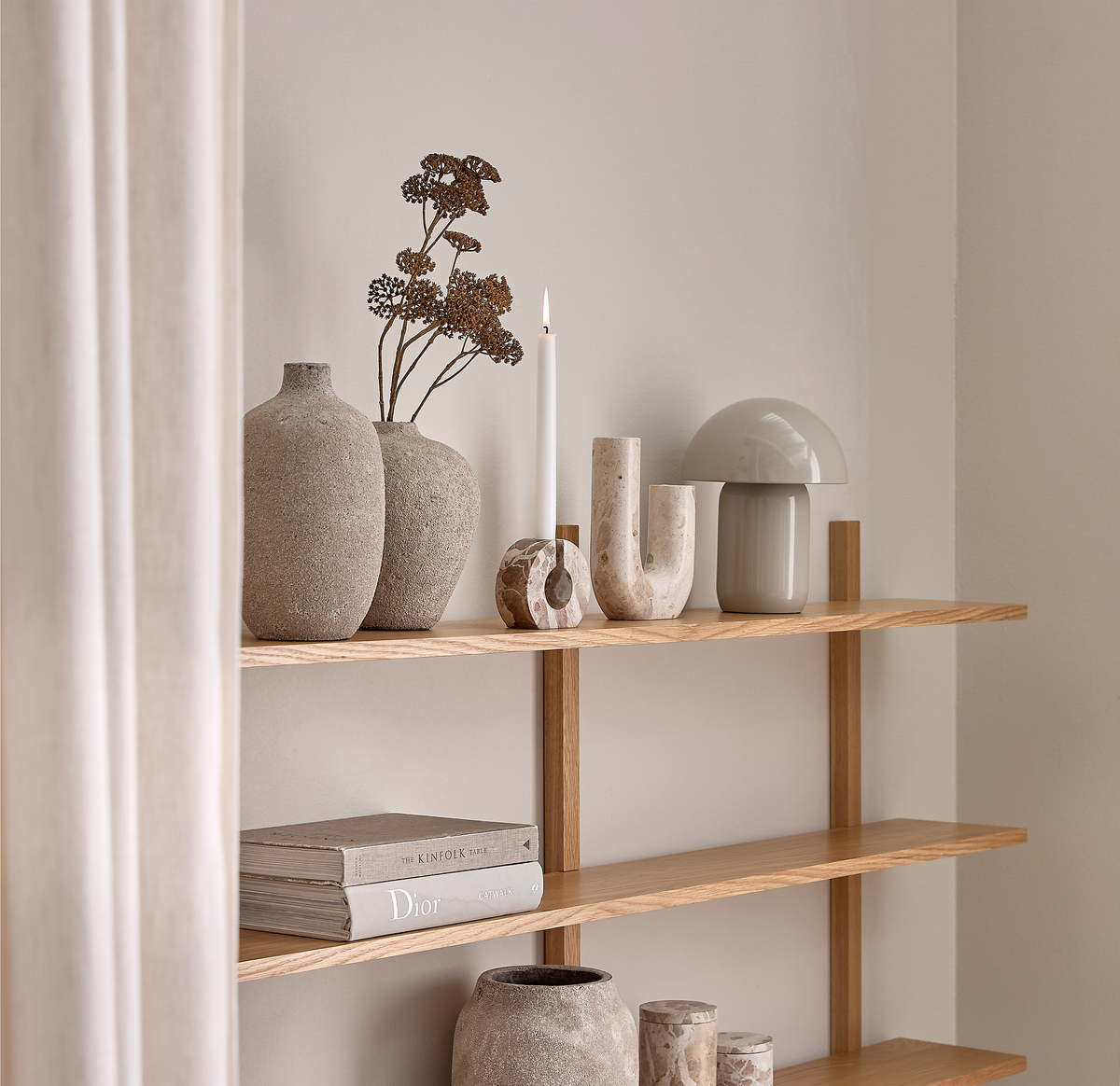 Lys træreol med keramikvaser, tørrede kviste, tændt stearinlys, skulpturelle stenobjekter, grå svampeformet bordlampe og bøgerne The Kinfolk og Dior i et lyst, minimalistisk hjem.