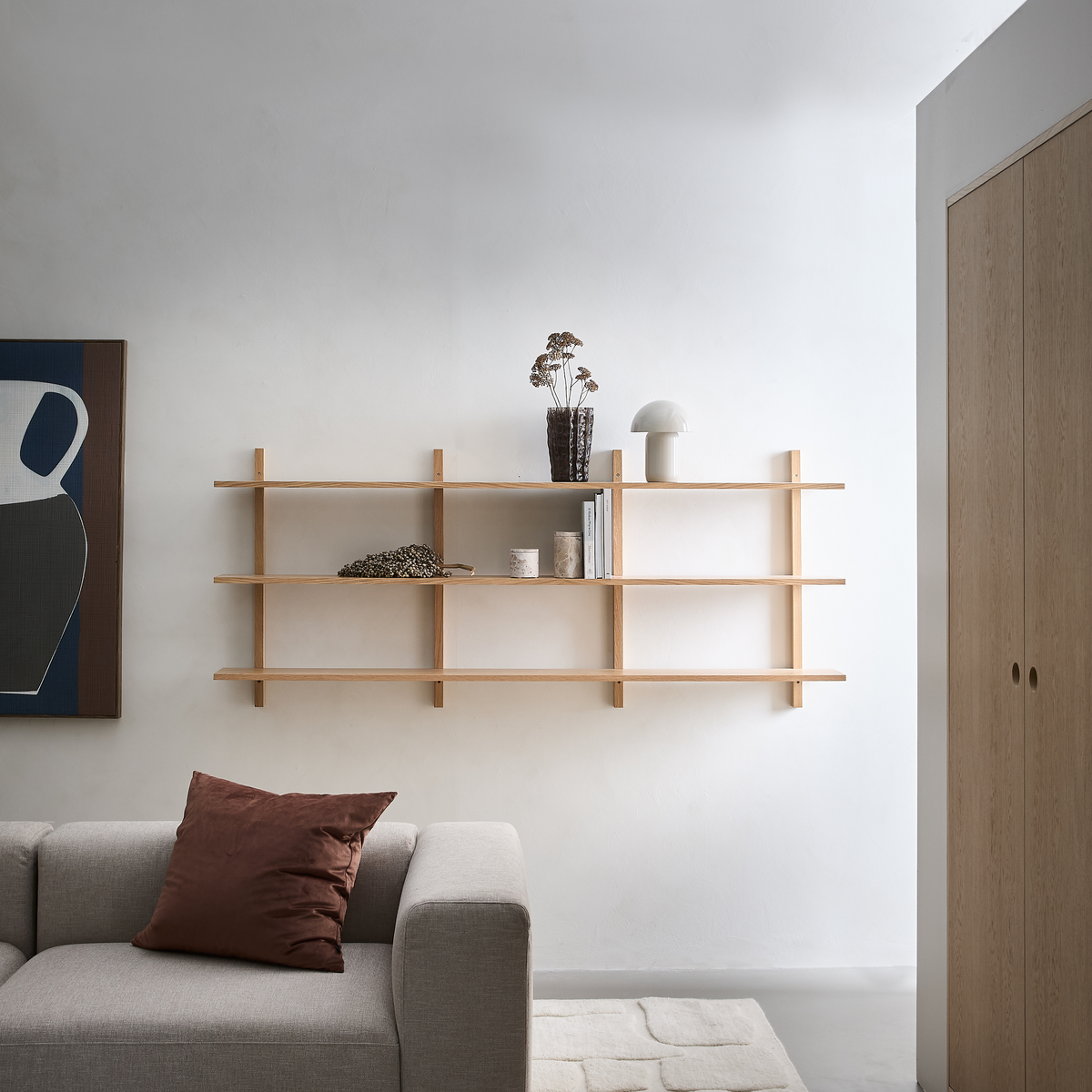 Lyst skandinavisk stue med vægmonteret hylde i lyst træ, neutral sofa med brun pude, kunst på væggen og dekorationer som bøger, vase med tørrede blomster og en lille hvid lampe.