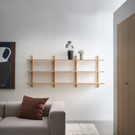 Lyst minimalistisk stue med vægmonteret træreol, tørrede blomster i vase og lille hvid bordlampe, grå sofa med brun pude, lyst tæppe og garderobeskab i træ.