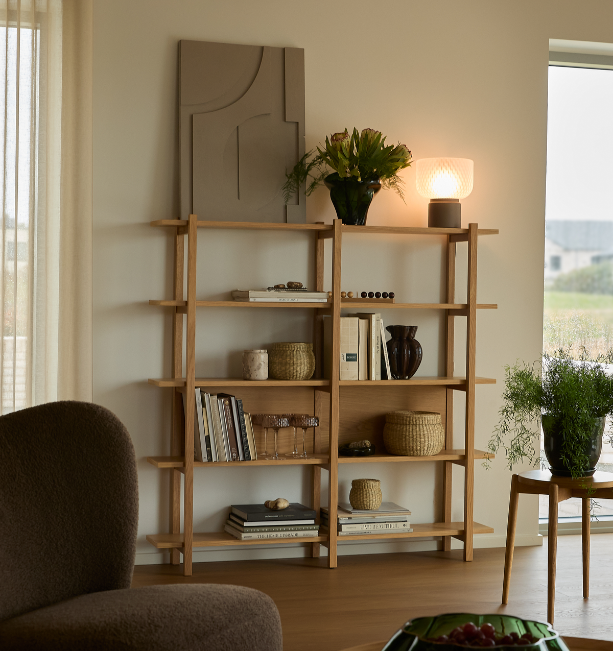 Lyst stue med reol i eg, bøger, kurve, vase med grønne planter og tænd bordlampe.