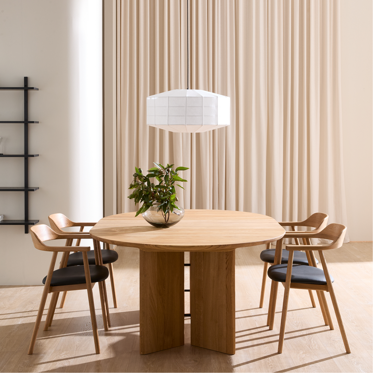 Minimalistisk spisestue med rundt egetræsbord og fire stole, hvid pendellampe, beige gardiner og sort væghylde; en glasvase med grønne grene står på bordet.
