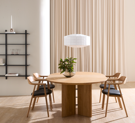 Minimalistisk spisestue med rundt egetræsbord og fire stole, hvid pendellampe, beige gardiner og sort væghylde; en glasvase med grønne grene står på bordet.