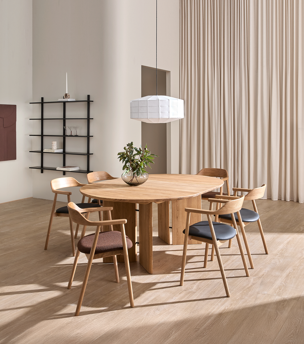 Minimalistisk spisestue med rundt egetræsbord, seks træstole, hvid pendel og glasvase med grønne grene; beige gardiner og sort hylde i baggrunden.