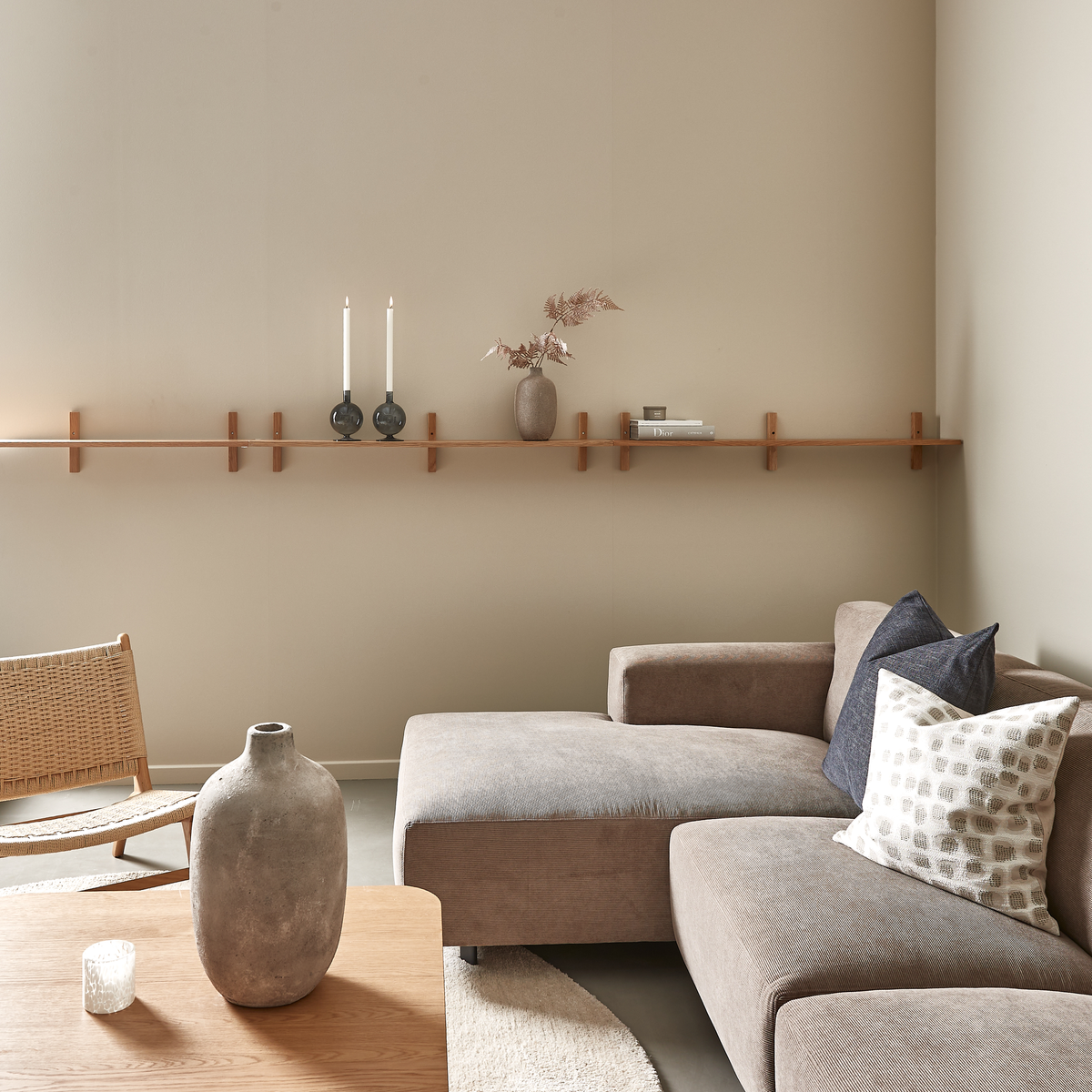 Beige stue med brungrå modulsofa, træbord med stor keramikvase og lang vægmonteret hylde med lysestager, vase med tørrede grene og bøger.
