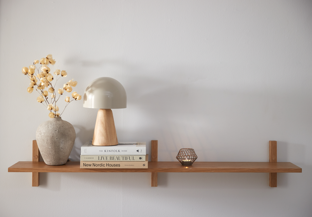 Træhylde på hvid væg med en rund betonvase med tørrede kviste, en beige bordlampe med træfod, tre bøger med titlerne The Kinfolk Home, Live Beautiful og New Nordic Houses samt en tænd lille lysestage i metal.