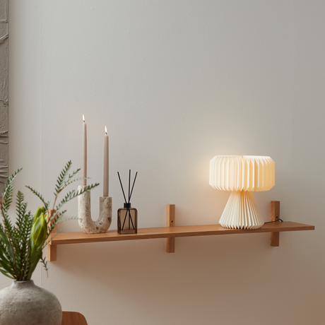 Vægmonteret træhylde med to stearinlys i stenholder, duftpinde i brun flaske og foldet bordlampe med varmt lys; minimalistisk skandinavisk indretning.
