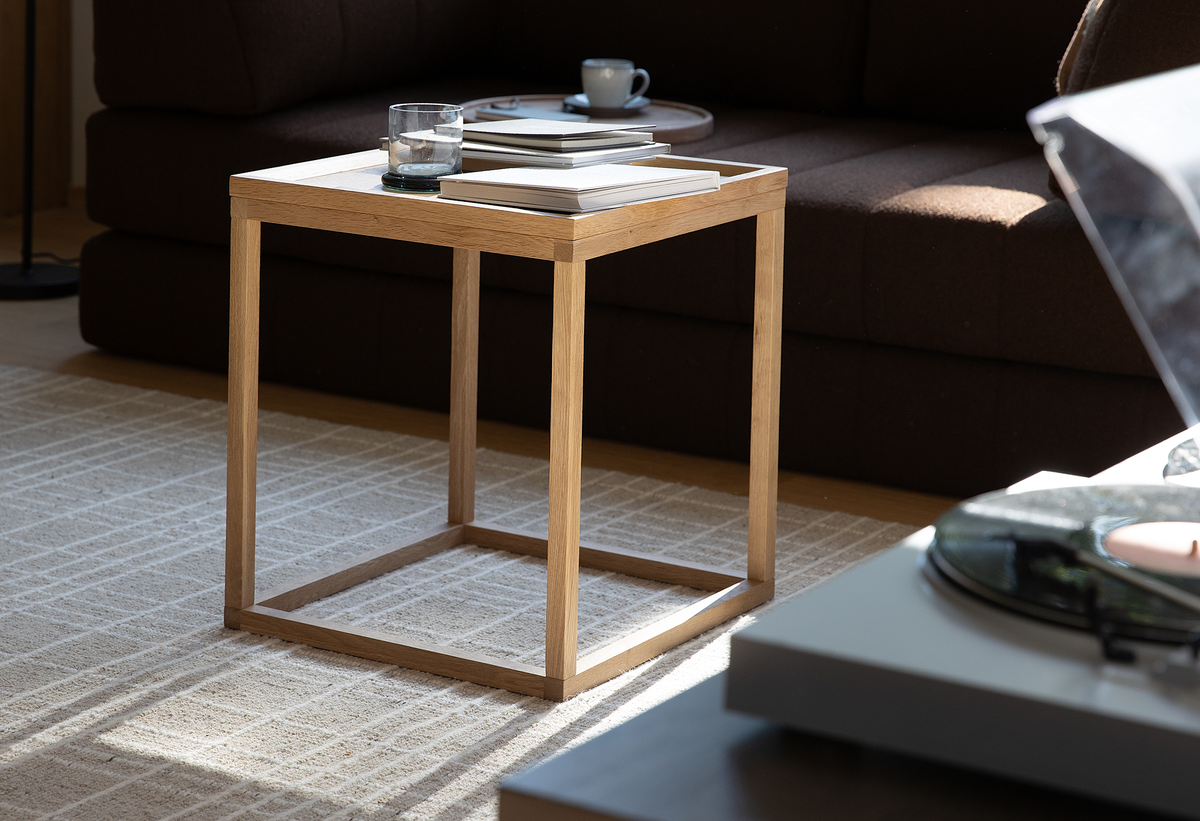 Kubisk sidebord i eg med bakke-top, med bøger og et glas, i en lys stue med sofa og tæppe.