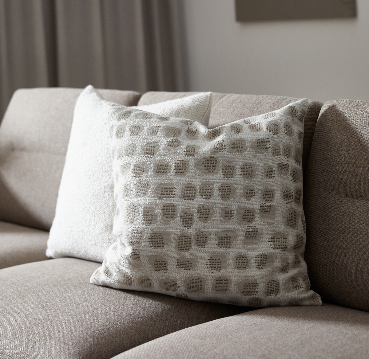 Beige sofa med to pyntepuder i neutrale nuancer; forreste pude har et lysbeige mønster med teksturerede prikker.