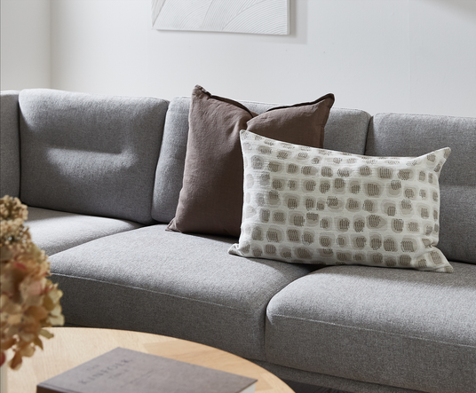Grå sofa med to pyntepuder – en brun og en beige med mønster – i et lyst stuemiljø med træbord og tørrede blomster.