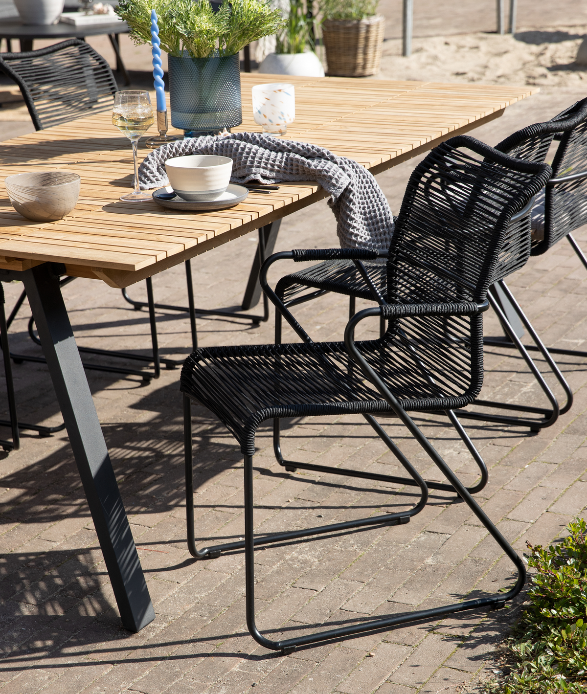 Terrasse med havebord i lyst træ og sorte rebstole; på bordet står en skål med underkop, vinglas, vase med grønne planter, lys og et gråt plaid.