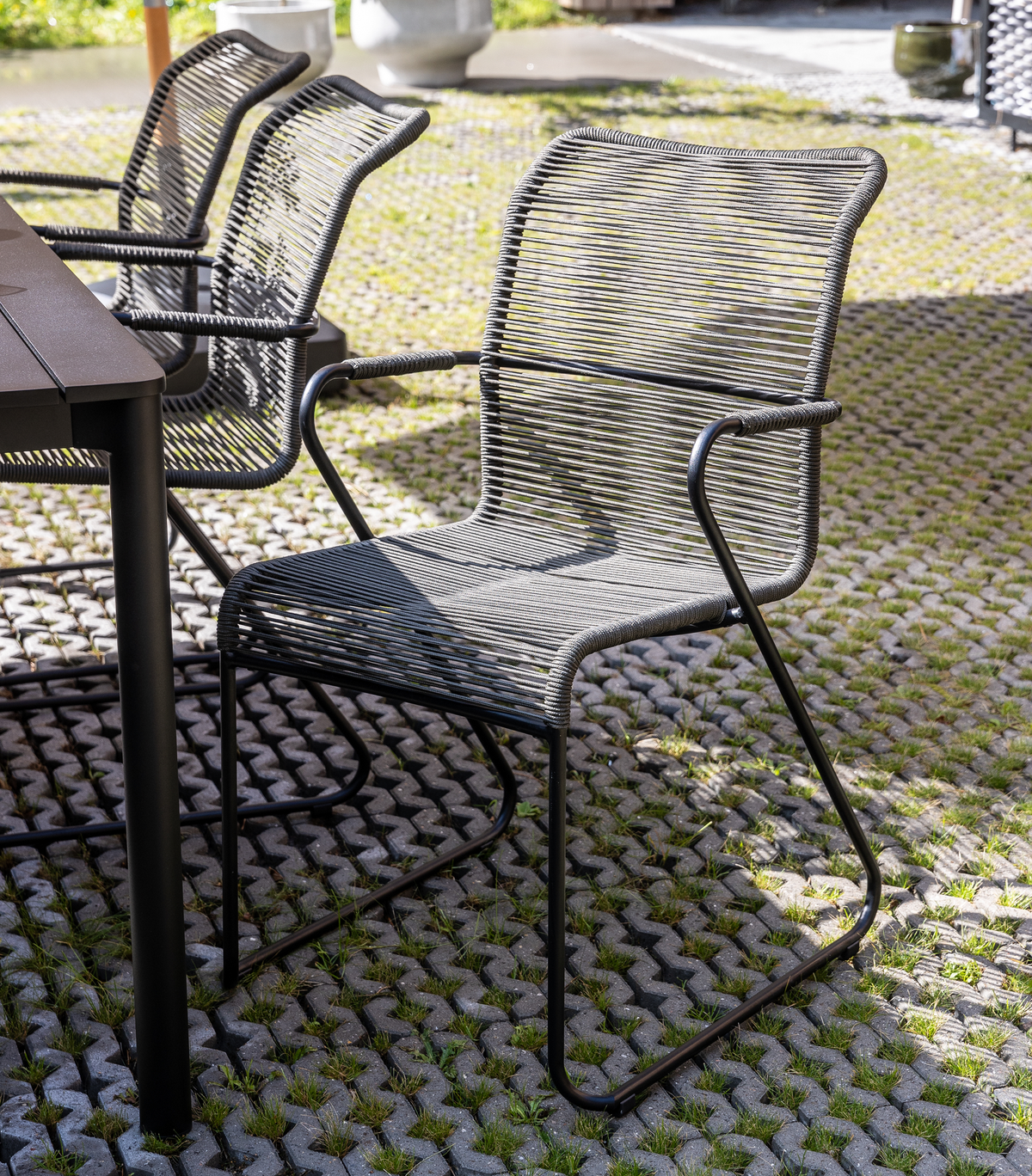 Moderne udendørs spisebordsstol i grå snor og sort metal ved siden af bord på brolagt terrasse i dagslys.