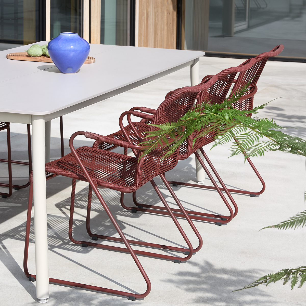 Udendørs terrasse med lysegråt spisebord, tre røde fletstole i metal og en blå vase på en bakke i sollys.
